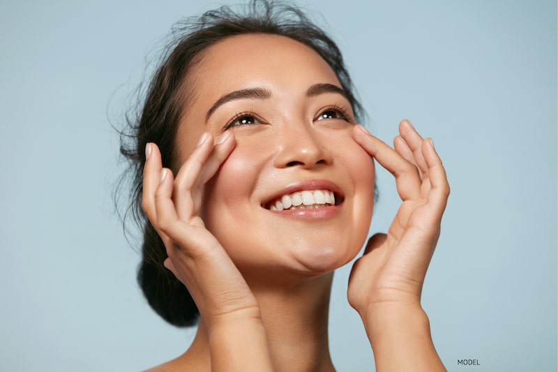 Young, Asian woman enjoying the effects of a youthful-looking face against a light blue backdrop.